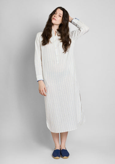 A woman wearing a long, white linen button-down kaftan with blue stripes and blue piping details on a plain grey background.