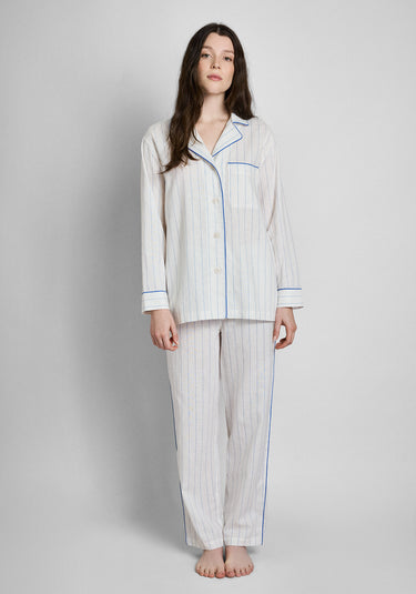 A woman wearing the Margaret Linen Pajama set, a white linen long sleeve and pants pajama set with blue stripes, blue piping details. Also features a chest pocket.