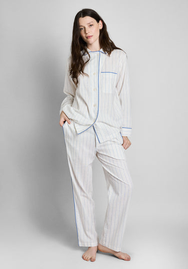 A woman wearing the Margaret Linen Pajama set, a white linen long sleeve and pants pajama set with blue stripes, blue piping details. Also features a chest pocket.