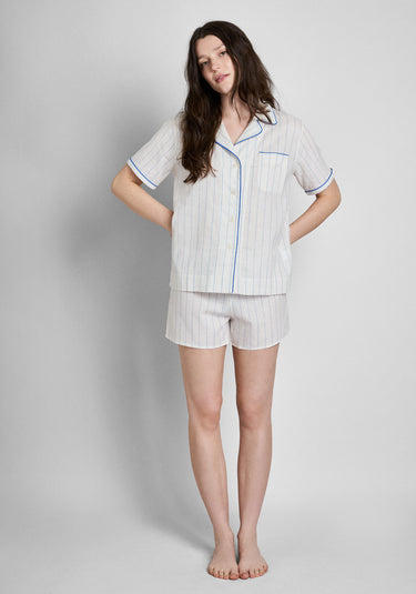 A woman wearing a short sleeve white linen pajama shirt with blue stripes and blue piping details and matching shorts on a plain grey background.