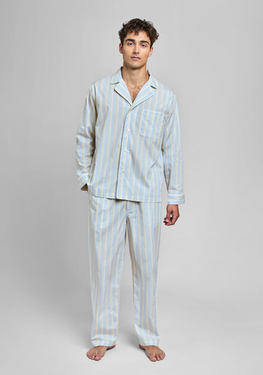 Man standin g wearing a ight blue pajama set with cream and tan stripes and white piping on a light grey background.