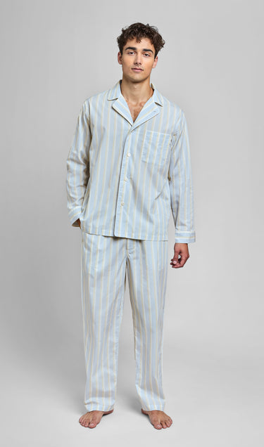 Man standin g wearing a ight blue pajama set with cream and tan stripes and white piping on a light grey background.
