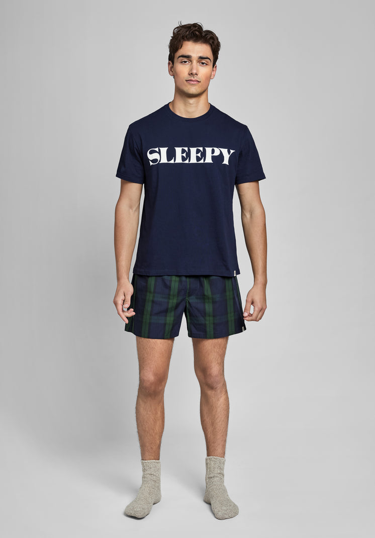 Man wearing a navy blue t-shirt with 'SLEEPY' text and navy and green blackwatch plaid boxers on a gray background