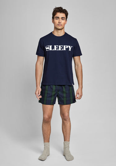 Man wearing a navy blue t-shirt with 'SLEEPY' text and navy and green blackwatch plaid boxers on a gray background