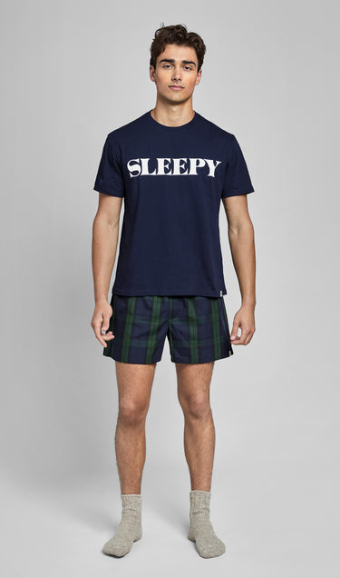 Man wearing a navy blue t-shirt with 'SLEEPY' text and navy and green blackwatch plaid boxers on a gray background