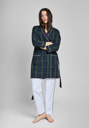 Woman wearing a green and navy blackwatch plaid robe with white piping over white pants on a gray background