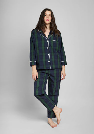 Woman wearing a green and navy blackwatch plaid pajama set with white piping details on a grey background