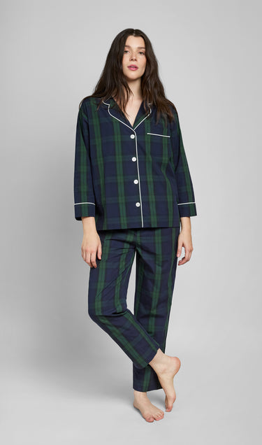 Woman wearing a green and navy blackwatch plaid pajama set with white piping details on a grey background