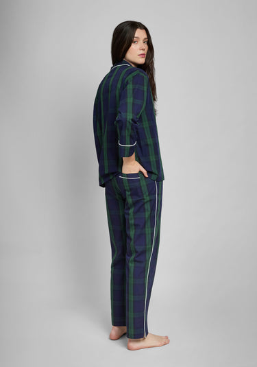 Woman wearing a green and navy blackwatch palid pajama set on a gray background. Her hand is in her back pocket.