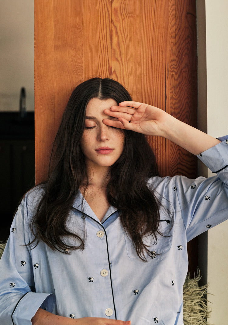 Woman in a light blue pajama top with small sheep patterns, sitting against a wooden wall.