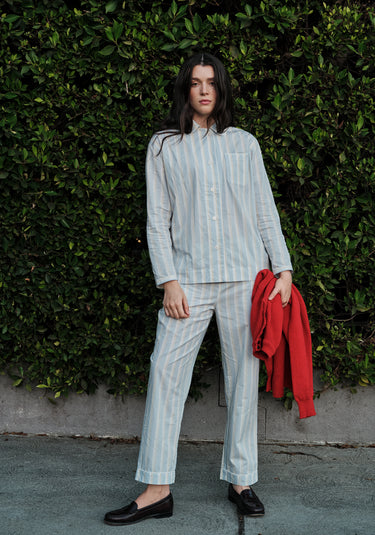 Woman wearing a light blue, sand, and white striped pajama set standing against a green hedge holding a red sweater.