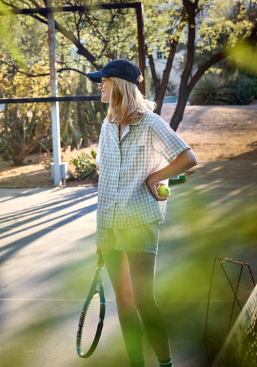 Woman holding a tennis ball and racket on a tennis court wearing a short sleeve and shorts pajama set in white and grey gingham.