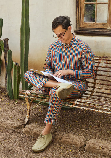 Man in colorful striped pajama set reading a book on a bench with cacti in the background