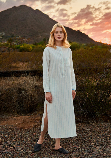 Woman in a long, white linen dress with blue stripes standing in a desert landscape with mountains and sunset.