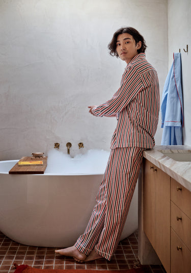 A man wearing a red, white, and blue striped pajama set stands in the bathroom next to a bathtub full of bubbles.