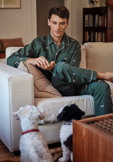 A man wearing green pajamas with dogs embroidered on them sits on a couch while his two dogs sit in front looking up at him. 
